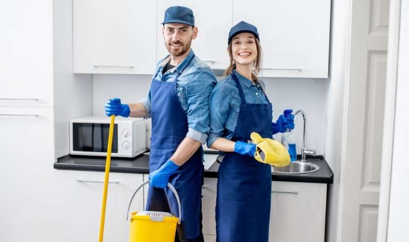 a young man and a woman standing inside a kitchen with a bucket, wiper and spray bottle