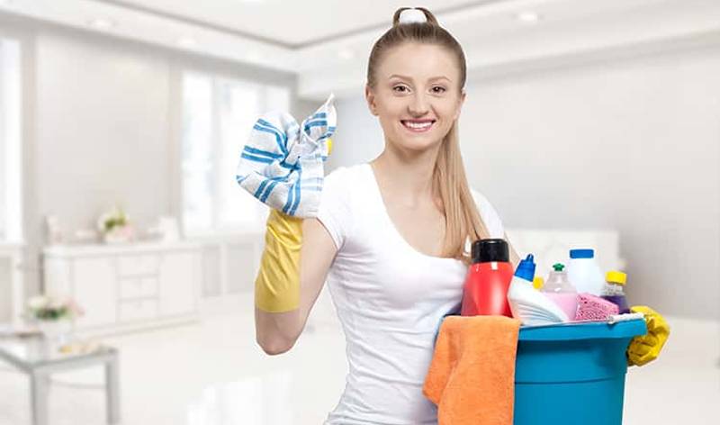 Woman in white t-shirt and yellow gloves holding a basket in one hand and cloth in another hand.