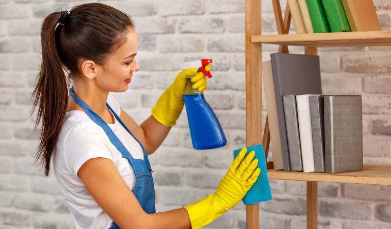 Woman in white-blue dress and yellow gloves holding a blue bottle and green scrubber in her hand