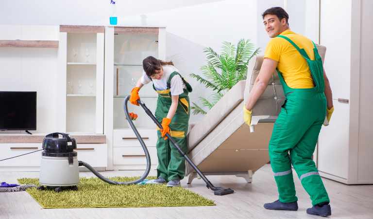 Man is holding a sofa and woman is vacuuming with vacuum cleaner.