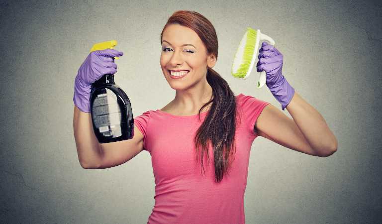 Woman in red top and purple gloves holding a black bottle and yellow brush in her hand.