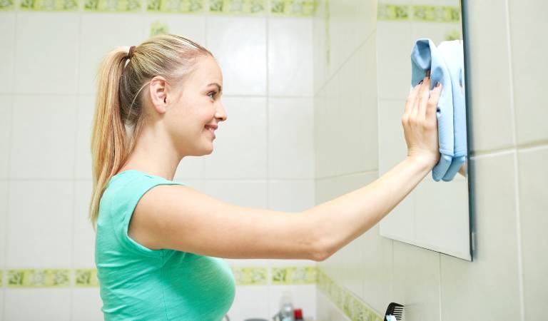 Smiling lady in blue top cleaning mirror inside her bathroom
