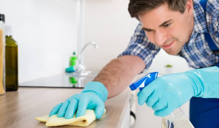 A man in check shirt and blue gloves holding a bottle and scrubbing a table with yellow cloth.