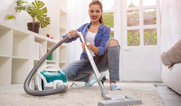Woman in check shirt and jeans vacuuming carpet inside her living room.