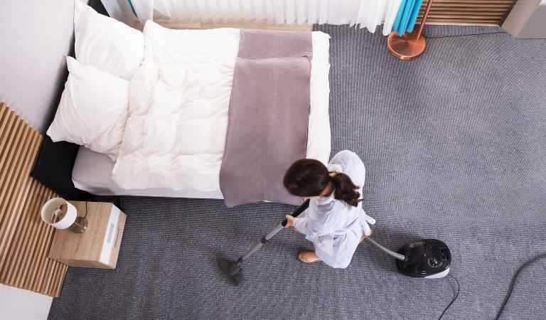 Woman in white uniform vacuuming carpet inside a bedroom.