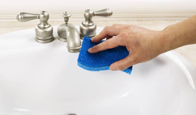 Hand with sponge cleaning bathroom sink faucet