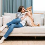 Woman in check shirt and blue jeans playing with her pet dog on sofa