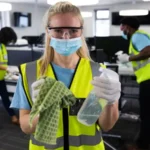 Woman wearing hi her vest, gloves, safety glasses and face mask, cleaning and sanitizing office with colleagues in the background