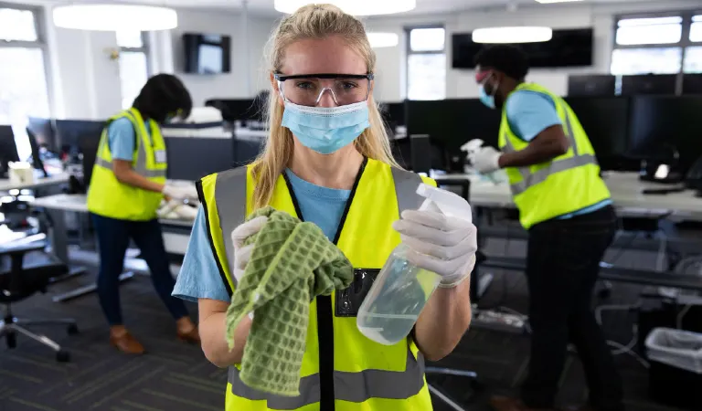 Woman wearing hi her vest, gloves, safety glasses and face mask, cleaning and sanitizing office with colleagues in the background