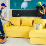 Young man with vacuum cleaner washing with vapor yellow sofa and beautiful woman wipes cuisine furniture.
